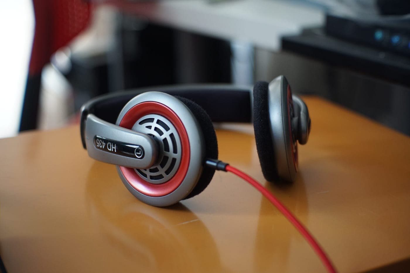 Close up image of a pair of over ear headphones with red accents and a red cable placed on a metal surface. The background is blurred, showing parts of an office interior